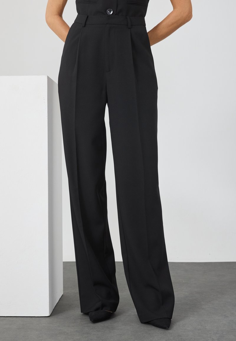 Femme portant un pantalon noir taille haute à jambes larges avec plis, debout les mains dans le dos devant un fond uni.