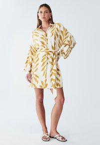 Abito camicia in tessuto bianco con motivi a foglia gialla. Presenta una vita con cintura e maniche lunghe e svasate. Indossato con sandali bianchi.