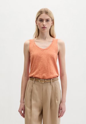 Female model with long blonde hair wearing an orange sleeveless top tucked into beige high-waisted pants with a slim belt, standing against white.