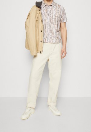 Chemise à manches courtes avec un motif beige et marron, associée à un pantalon couleur crème et une veste beige. Des baskets blanches complètent la tenue.