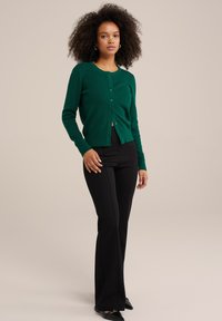 Zaļš adīts kardigāns ar teksturētu rakstu, apaļu izgriezumu un pogu aizdari, kombinēts ar melnām plati apgrieztām biksēm un plakanām kurpēm.