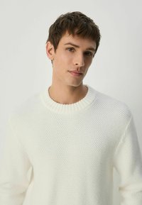 White textured sweater with a ribbed crew neckline and relaxed fit. The fabric features a patterned knit, offering a soft appearance.