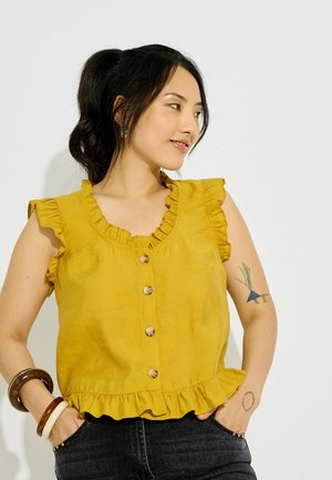 Femme aux cheveux foncés attachés en queue de cheval, portant un haut jaune sans manches avec des volants et des boutons, un pantalon noir et des bracelets à son poignet gauche.