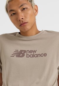 Camiseta de algodón beige con cuello redondo, que presenta un logo impreso de "new balance" en marrón oscuro, con un diseño de rayas horizontales en el pecho.