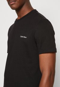 Sort crewneck t-shirt lavet af bomuld, der har et hvidt broderet Calvin Klein-logo på venstre bryst. Korte ærmer, glat tekstur.