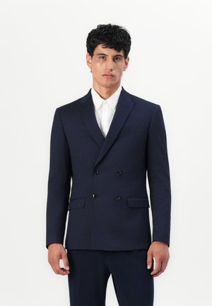 Μαρίνας ναυτικού σακάκι διπλής όψης με κανονικό πέτο, μαύρα κουμπιά, δύο μπροστινές τσέπες και λεία υφή υφάσματος, το οποίο συνδυάζεται με ένα λευκό πουκάμισο.