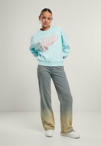 Felpa azzurro chiaro con logo "Kani" in rosa, abbinata a jeans a gamba larga con una sfumatura gradiente dal blu al beige, indossata con sneakers bianche.