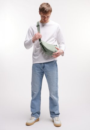 Jonge man met wit shirt met lange mouwen, lichtblauwe spijkerbroek en sneakers die een lichtgroene schoudertas verstelt tegen een effen witte achtergrond.