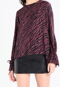 Blouse à motifs zèbre dans des tons marron et noir, avec des manches longues et des détails à nouer aux poignets, assortie à une mini jupe noire texturée.