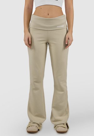 Donna che indossa pantaloni beige a gamba larga con vita pieghevole e pantofole beige, in piedi contro uno sfondo chiaro e uniforme.