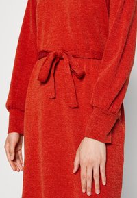 Vestido rojo texturizado con mangas largas y un lazo en la cintura. La tela parece suave, con un patrón sutil y un ajuste ligeramente holgado en los puños.