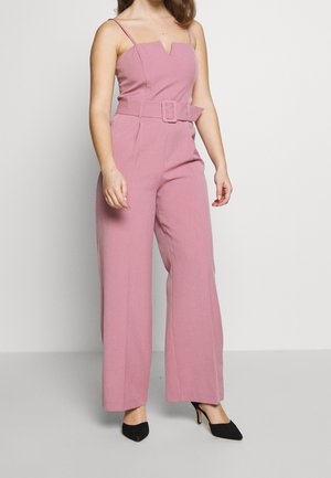 Femme portant une combinaison pantalon large couleur mauve avec bretelles fines et ceinture assortie, accompagnée de talons pointus noirs, se tenant devant un fond uni.