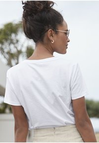 Weißes Baumwoll-T-Shirt mit kurzen Ärmeln, lässiger Passform und rundem Halsausschnitt. Modell von hinten betrachtet, kombiniert mit einem hellen Rock.