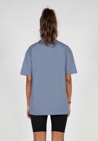 Blå bomull T-skjorte med korte ermer, en avslappet passform og rund halslinning, brukt med svarte leggings; enkel bakview.