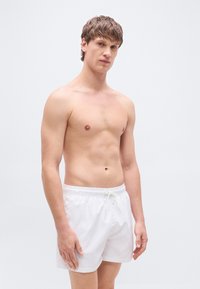 Λευκό μαγιό βερμούδα με ελαστική μέση και κορδόνι. Λείο ύφασμα, μήκος μέχρι το μέσο του μηρού, χωρίς εμφανή σχέδια ή διακοσμήσεις.