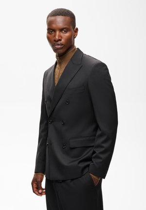Homme debout portant une veste de costume noire croisée sur un pull à col roulé marron sur un fond blanc uni.