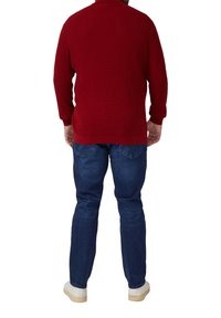 Rdzawo-czerwony sweter z dzianiny o żebrowanej fakturze, z długimi rękawami i wysokim kołnierzem, w połączeniu z niebieskimi jeansami i białymi trampkami.