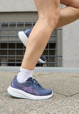 Osoba biegająca na zewnątrz, nosząc niebiesko-różowe buty do biegania ASICS i białe skarpetki do kostek na brukowanym miejskim chodniku.
