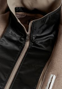 Schwarze Jacke mit Reißverschluss und glattem Satin-Innenfutter, beigefarbener Außenstoff und kontrastierende Reißverschlüsse. Mit hohem Kragen und seitlichem Reißverschlussfach.