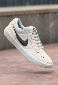 Adidași Nike SB, cu un superior din suede gri deschis și crem, swoosh negru și vârful perforat, concepuți pentru skateboard.