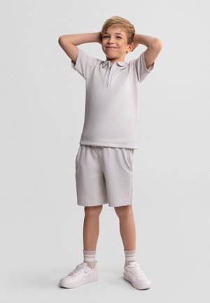 Młody chłopiec z krótkimi blond włosami, w jasnoszarej koszulce polo na suwak i dopasowanych szortach, z rękami założonymi za głowę, stoi na jednolitym tle.