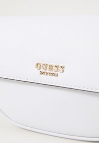 Borsa in finta pelle bianca con logo "GUESS" dorato, design elegante, dettagli cuciti e una sottile rifinitura nera lungo i bordi.