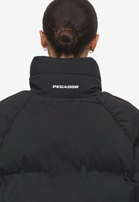 Svart pufferjacka med hög krage, som har en framträdande logotyp "PEGADOR" på baksidan. Slät textur och strömlinjeformad design.