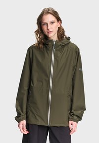 Νεαρή γυναίκα που φοράει πράσινο μπουφάν με κουκούλα, εμπρός φερμουάρ και τσέπες με φερμουάρ, στέκεται μπροστά σε ένα απλό φωτεινό φόντο.