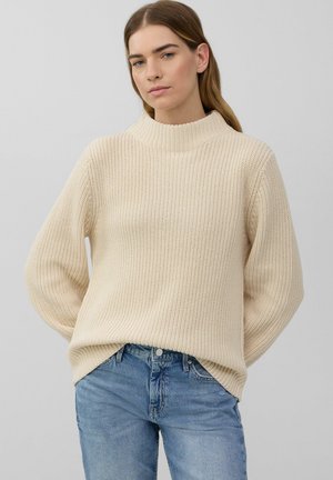Maglione chunky in beige chiaro, con colletto alto, vestibilità ampia e texture a costine. Abbinato a jeans in denim azzurro.