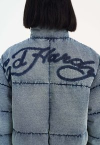 Denim pufferjakke med en blå quiltet tekstur, der har mørkeblå graffiti-agtig skrifttype på ryggen, høj krave og syet sømme.