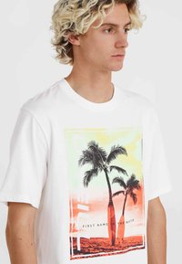 Camiseta de algodón blanca con un gráfico multicolor de palmeras y tablas de surf, con un contorno rectangular y un texto superpuesto.