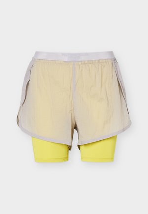Béžové voľné vonkajšie šortky so žltými priliehavými vnútornými šortkami viditeľnými pod nimi, obidva s elastickými pásmi, zobrazené na bielom pozadí.