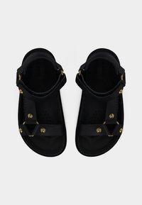 Sandalias negras de cuero con correas en el tobillo y tachuelas doradas, mostradas desde arriba sobre un fondo blanco liso.