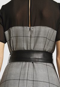 Haut noir avec tissu transparent, fermeture éclair sans couture à l'arrière ; le corps présente une jupe à carreaux noir et blanc avec une large ceinture noire.