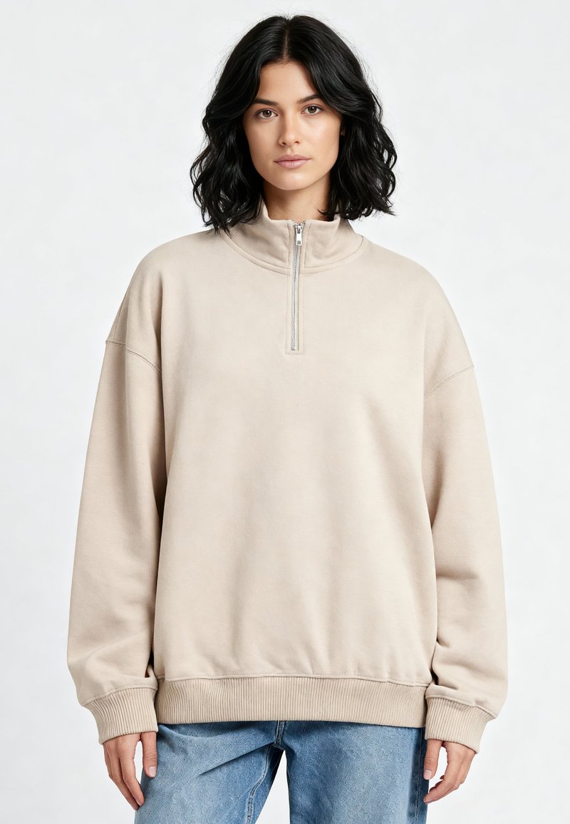 Femme aux cheveux foncés mi-longs portant un sweat-shirt beige à fermeture éclair quart et un jean bleu clair, debout devant un fond uni.