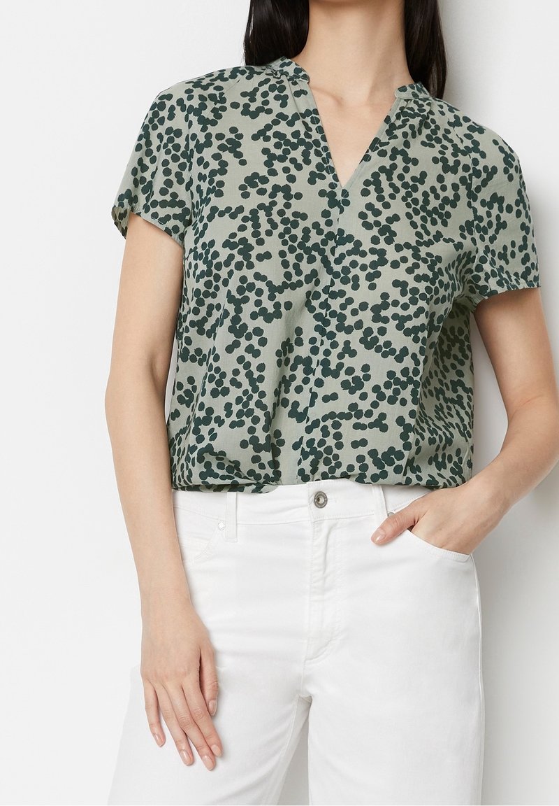 Blouse à manches courtes en tissu léger, avec un fond vert et des motifs circulaires vert foncé, col en V et coupe décontractée.