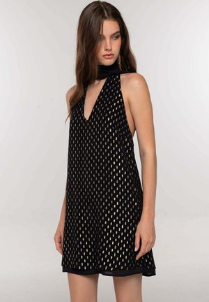 Jeune femme portant une robe sans manches noire avec des motifs ovales dorés et un décolleté en V profond, regardant à sa gauche sur un fond uni.