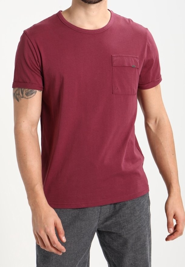 Homme portant un T-shirt bordeaux à manches courtes avec une poche poitrine et un pantalon gris, debout devant un fond blanc uni.