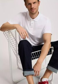 Biała bawełniana koszulka polo z kołnierzykiem, zestawiona z ciemnoniebieskimi dżinsami i podwiniętymi rękawami. Modelka siedzi na białym, drucianym krześle, w czerwono-białych sneakersach.