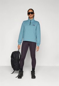 Világoskék polár pulóver cipzáros gallérral, fekete leggings, fekete túracipő és fekete hátizsák. Simább textúra és sportos fazon.