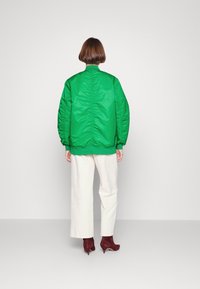 Giubbotto imbottito verde con una finitura tessurizzata, caratterizzato da dettagli arricciati lungo la schiena, abbinato a pantaloni bianchi a gamba larga e stivali rosso scuro.