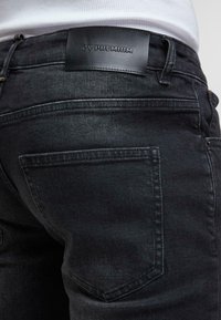 Gros plan d'un jean en denim noir montrant une poche arrière et une étiquette en cuir noir avec "ZV PREMIUM" embossé au-dessus de la poche.