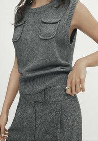 Haut en tricot sans manches gris avec un col côtelé, deux poches poitrine et une finition texturée. Assorti à un pantalon gris taille haute.