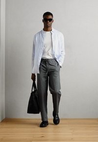 Jachetă deschisă la culoare, cu nasturi, tricou alb, pantaloni gri, pantofi negri și ochelari de soare. Geantă neagră tip tote. Design simplu, modern.