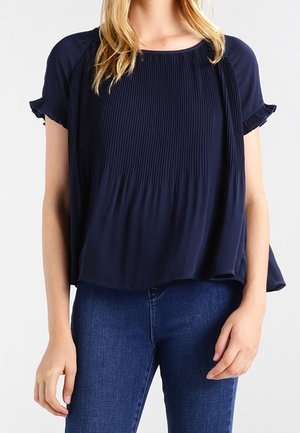 Kobieta ubrana w granatową plisowaną bluzkę z krótkim rękawem w połączeniu z dopasowanymi ciemnoniebieskimi dżinsami na neutralnym tle.