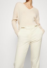 Pull en maille beige avec un col en V et des manches côtelées, assorti à un pantalon large crème clair avec une ceinture chaîne dorée en accent.