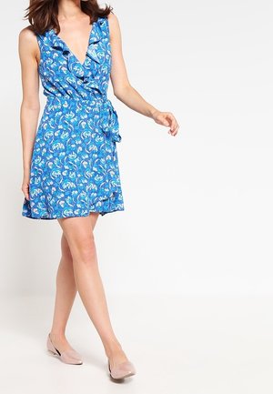 Kvinde iført en ærmeløs blå floral slå-om-kjole med flæser, kombineret med lyserøde flade sko, gående med den ene arm let hævet.