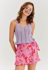 Levanduľový pletený crop top s volánovým lemom skombinovaný s ružovou kvetinovou obálkovou sukňou s volánmi a viazaním. K dispozícii sú aj okrúhle slnečné okuliare.