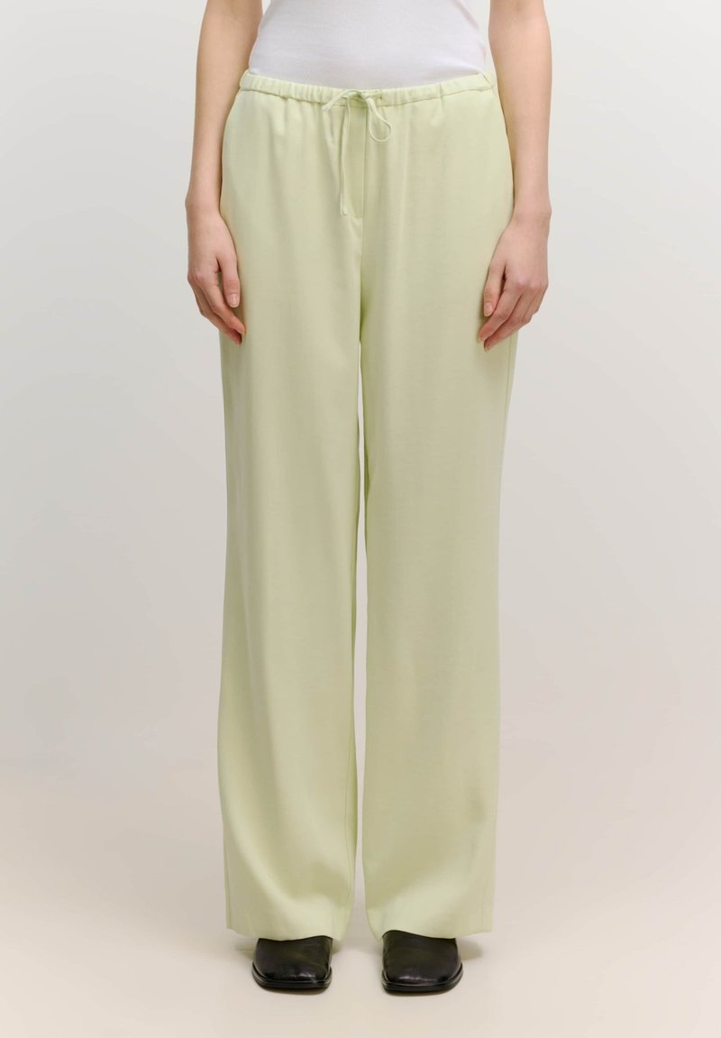 Lichtgroene, wijde broek met een elastische taille en trekkoord. Glad stofoppervlak, gedragen met donkere schoenen, weergegeven tegen een neutrale achtergrond.