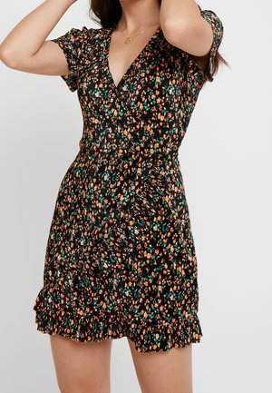 Femme portant une robe portefeuille courte noire à fleurs orange et vertes avec un ourlet à volants, posant les bras levés.